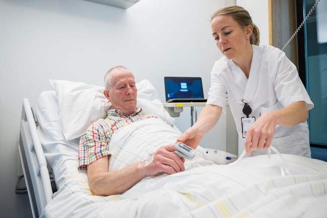 Kvinnelig sykepleier i hvit uniform kobler oksygenmåler på mannlig pasient som ligger i sykehusseng.
