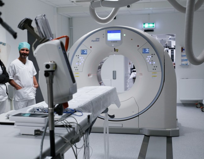 Hvit CT- og røntgenmaskin. En mann står på venstre side av maskinen. Foto. 