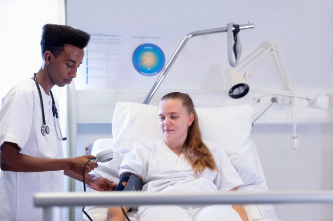 Mannlig student i hvit uniform måler blodtrykk på kvinnelig student i uniform som ligger i sykehusseng