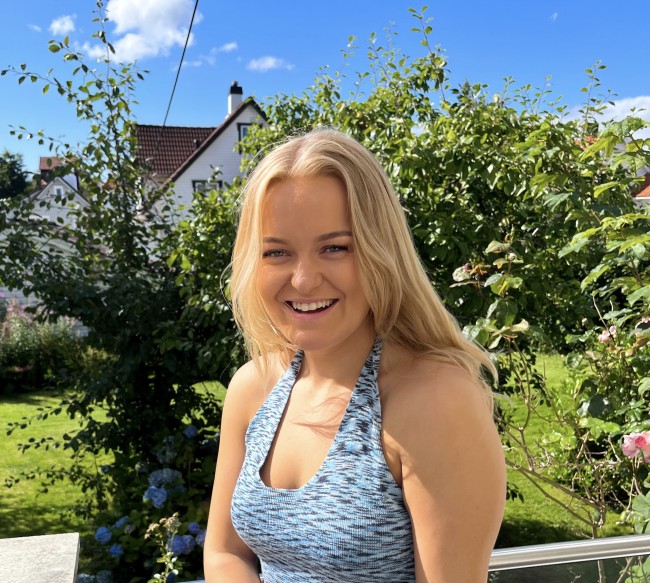 Kvinne som smiler mot kamera. Grønn hage i bakgrunnen. Foto