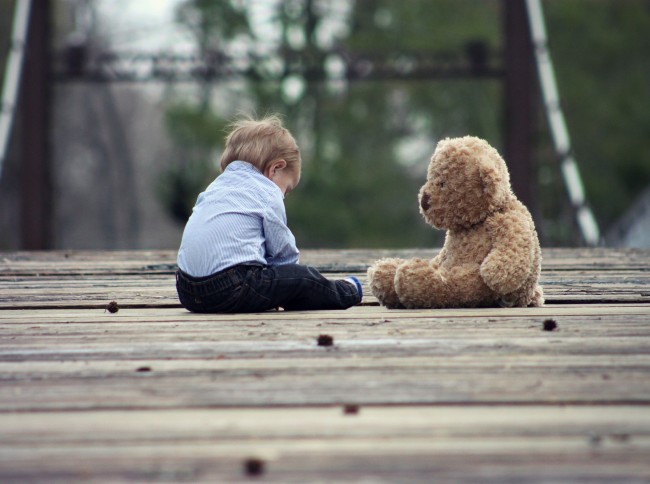 En baby i mørk bukse og blåstripete skjorte siter på et tregulv ute med en teddybjørn like ovenfor seg. Foto. 