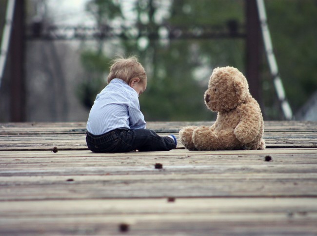 En baby i mørk bukse og blåstripete skjorte siter på et tregulv ute med en teddybjørn like ovenfor seg. Foto.