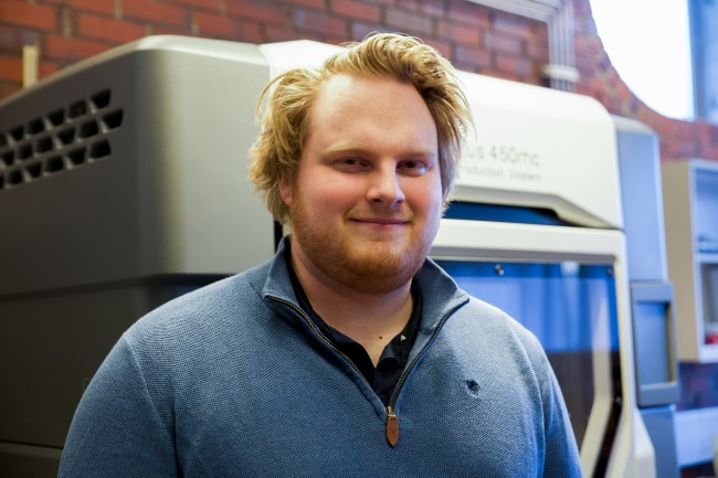 Portrettbilde av mann med blå genser som smiler til kamera. I bakgrunnen er en 3D-printer. Foto.