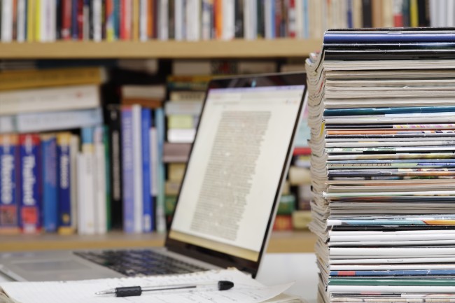En PC står på et bord med en bunke tidsskrift i forgrunnen og en bokhylle med bøker i bakgrunnen