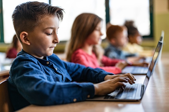 Elever i tiårsalderen sitter på en rekke og skriver på hver sin pc. Gutt fremst.