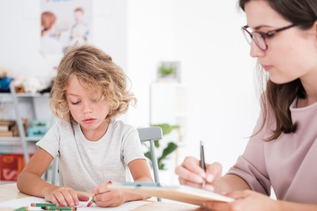 Et barn og en voksen kvinne sitter ved et bord. Barnet tegner og kvinnen skriver noe på et skjema.