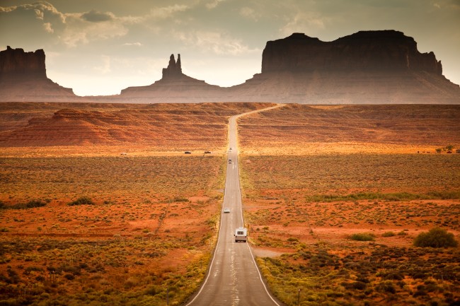 åpent landskap med kjørende bil på bilvei mot horisont med fjell og solnedgang