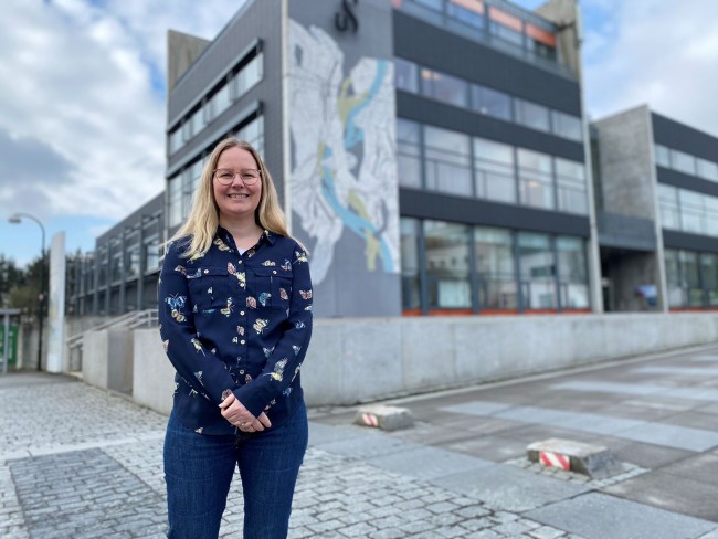 Markedsrådgiver ved UiS Susanna King står foran Arne Rettedals hus på campus Ullandhaug, en høy bygning i grått og oransje.