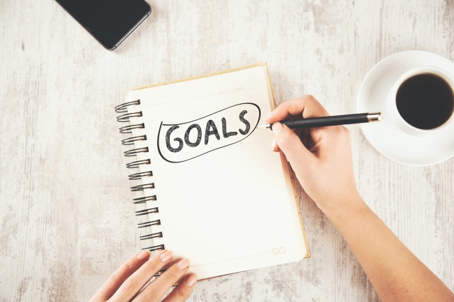 Hånd som skriver ordet "Goals" (mål) på notatblokk med kaffekopp ved siden av.
