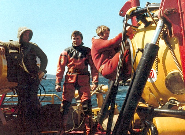 tre menn i dykkerdrakt på en oljeinstallasjon. En av dem er på vei inn i en dykkerklokke.