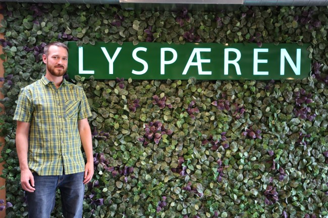 En person står foran en grønn vegg med blader og et skilt der det står Lyspæren.