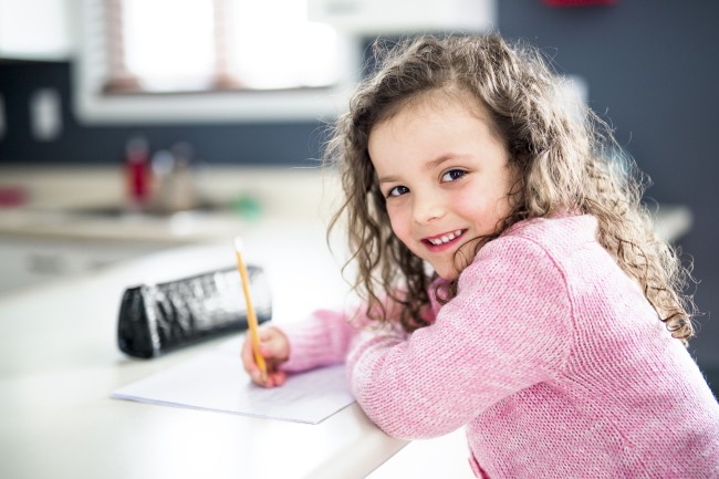 LIten jente skriver på ark og ser i kamera med et smil