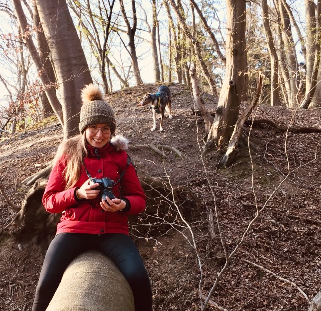 En person sitter ute i skogen og smiler mot kamera. I bakgrunnen er trær og en hund. Foto.