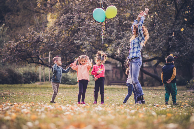 Fem barn og en voksen utendørs. Ett av barna holder to ballonger. Den voksne hopper høyt i været.