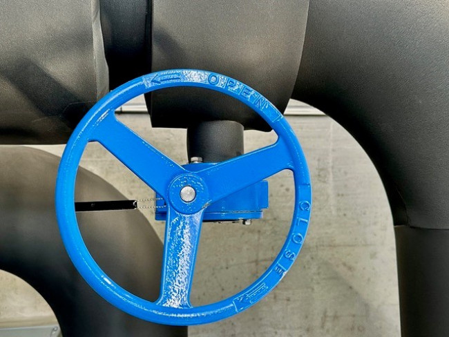 A blue valve wheel on a grey pipe with a grey concrete wall behind it
