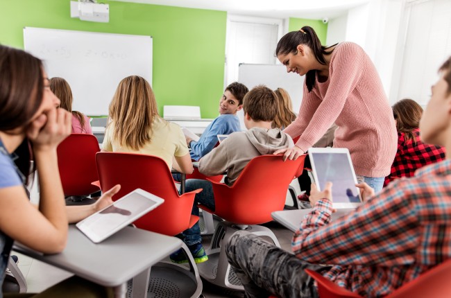 Klasseromssitausjon er lærer står ved siden av elev midt i klasserommet og snakker med ham. Elevene rundt følger med på samtalen.