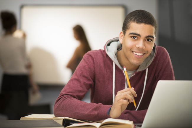Vgs-elev som ser rett i kamera og smiler. Sitter foran en skjerm, med en blyant i hånden og medelever i et klasserom i bakgrunnen.
