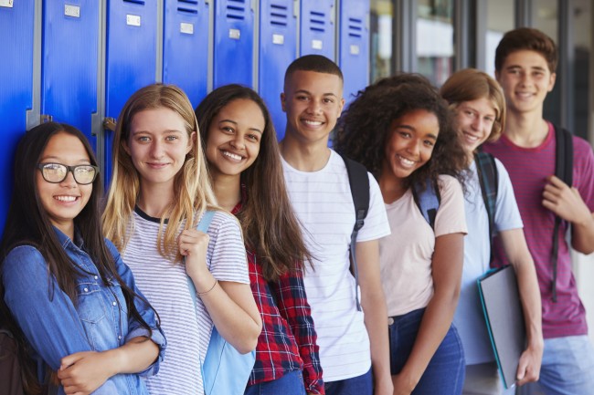 En gruppe smilende ungdommer står etter hverandre ved en skaprekke på en skole.